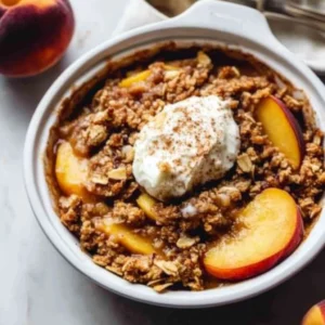 A white bowl filled with a generous serving of warm peach crisp, topped with a scoop of melting vanilla bean ice cream.